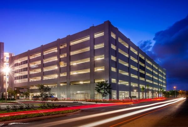 Exterior photo of the Miami Dade College Hialeah Campus parking garage. Evening photo with bright blue skies. 车库灯亮着. Vehicle lights create streaks on the adjacent street.
